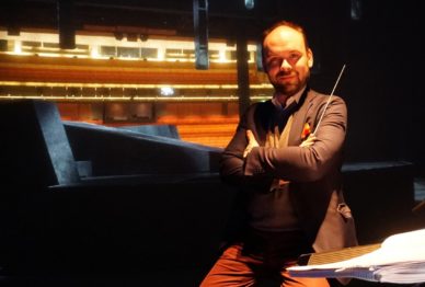 Mies istuu näyttämöllä partituurin äärellä kapellimestrain tahtipuikko kädessä