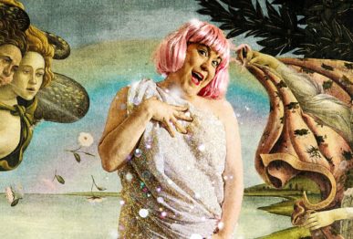 Vaaleanpunaperuukkinen nainen virnistää lakanaan kääriytyneenä simpukan sisällä seisoen